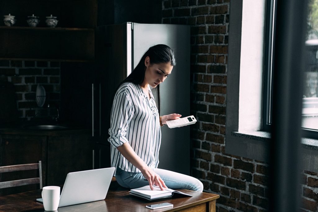 sad young woman with calculator counting tax on kitchen at home